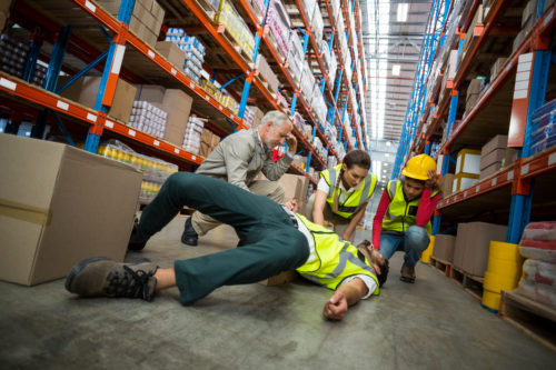 Man hurn and on floor in warehouse with three people kneeling by him to help