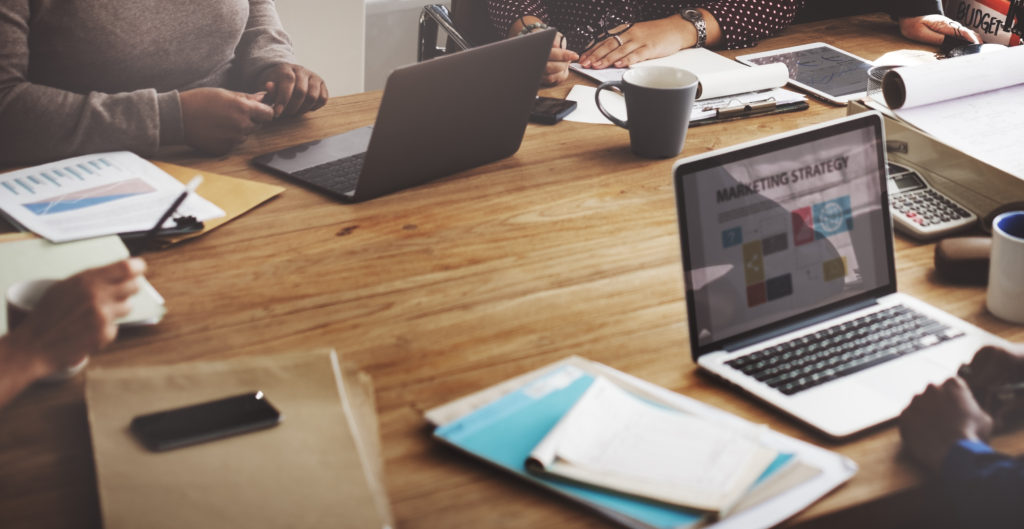 Professionals at a meeting with a laptop open on the table that says "Marketing Strategy" 