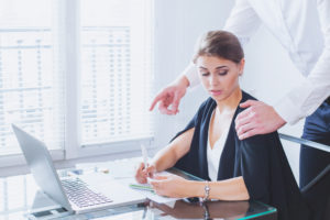 Man leaning over woman with a hand on her shoulder and another hand pointing to her computer while woman is visible uncomfortable