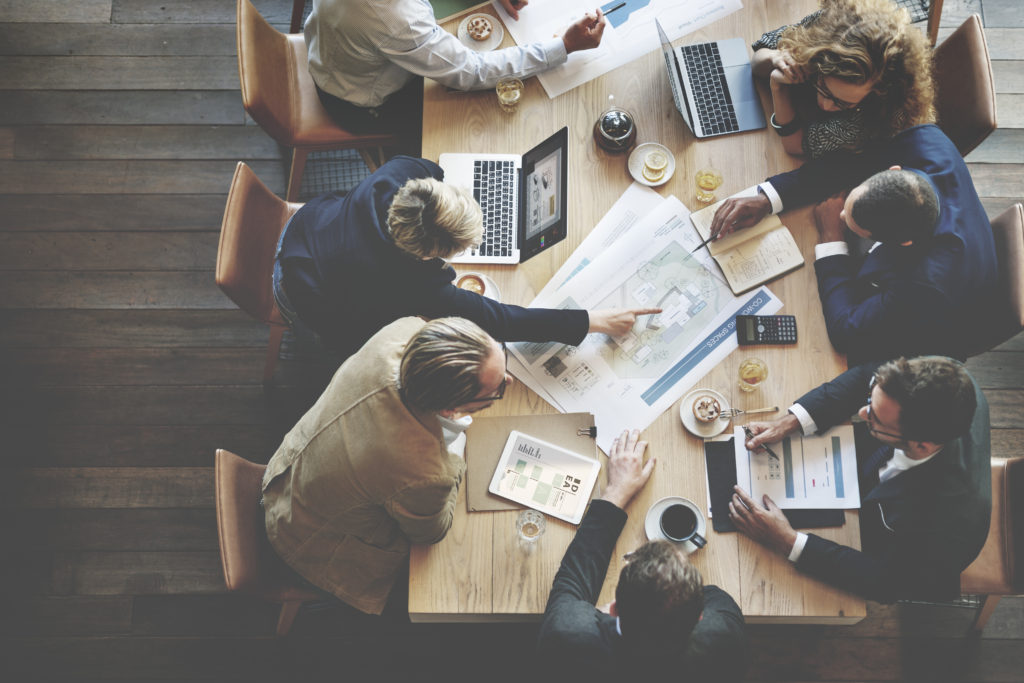 Professionals with laptops and notes at a table pointing at a diagram in the center of the table 